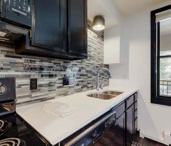 A modern kitchen with black appliances and cabinets, white countertops, a stainless steel sink, and a tiled backsplash; a window offers outdoor views.