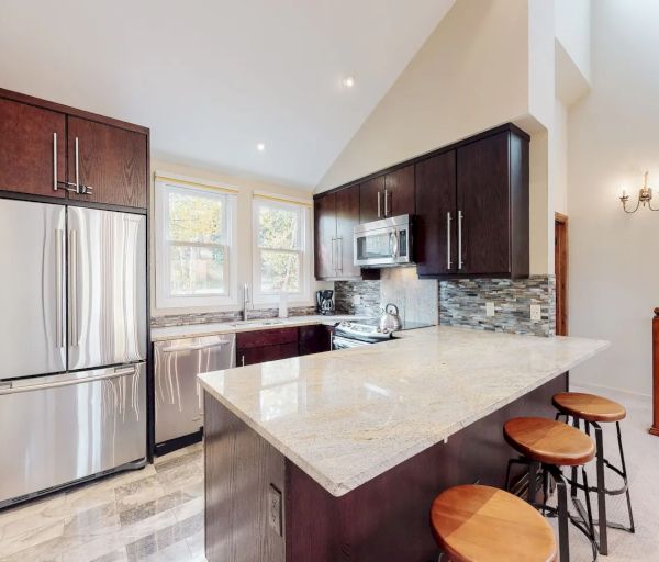 A modern kitchen with stainless steel appliances, dark brown cabinets, a white countertop, and three barstools.