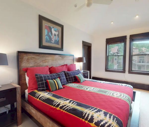 A modern bedroom with a large bed, vibrant red bedding, two nightstands with lamps, a ceiling fan, artwork, and two large windows.