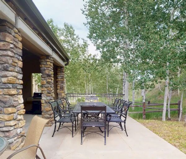 The image shows an outdoor patio area with a table, multiple chairs, stone pillars, and a view of a green landscape and trees in the background.