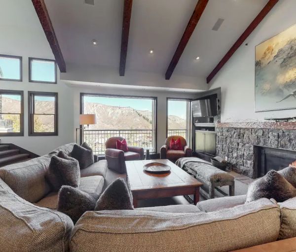 A cozy living room with a large sectional sofa, stone fireplace, mountain views through large windows, and wooden ceiling beams.