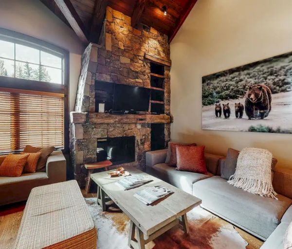 A cozy living room with a stone fireplace, large windows, sofas, and a bear-themed wall art.