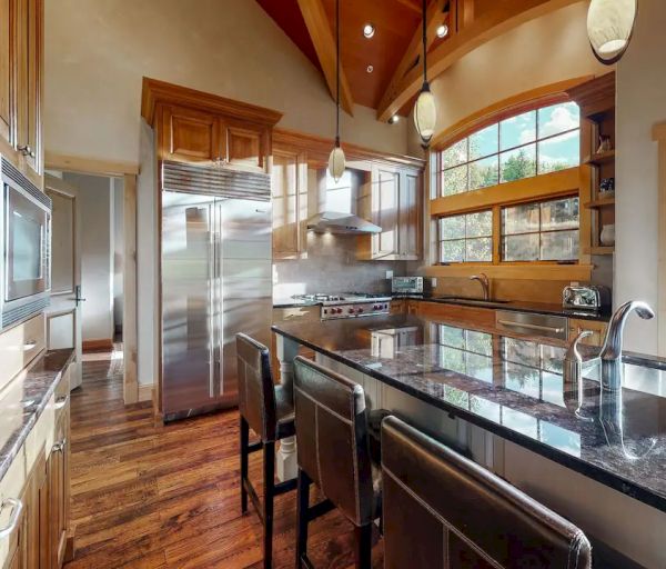A modern kitchen with wooden cabinets, granite countertops, stainless steel appliances, large windows, and high ceilings with pendant lighting.