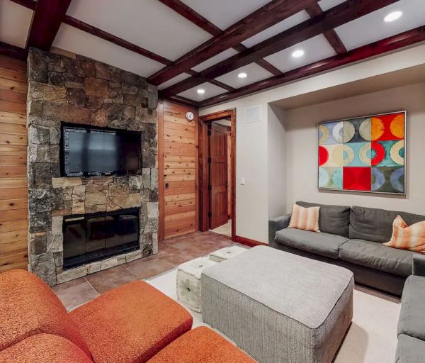 Cozy living room with stone fireplace, TV, modern artwork, and gray sofas with orange and beige accents. Wood beams and neutral decor.