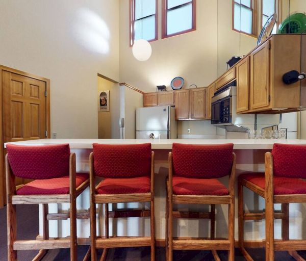 A kitchen with a high ceiling, four red cushioned bar stools at a counter, wooden cabinets, and stainless steel appliances.