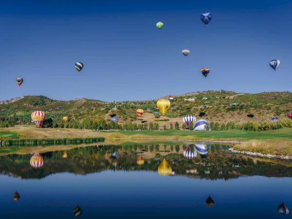 This image shows numerous colorful hot air balloons floating above a scenic landscape with a clear blue sky and a reflective lake below, ending the sentence.