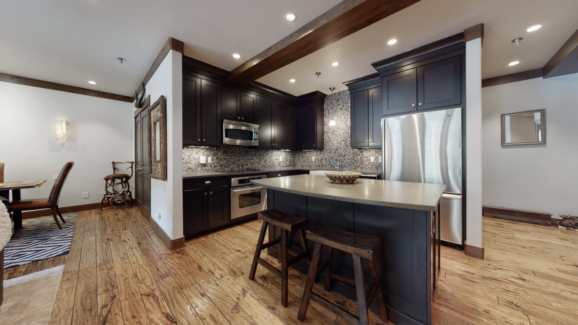A modern kitchen featuring dark cabinetry, stainless steel appliances, an island with stools, and a dining area to the side.