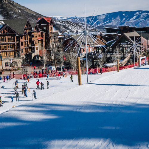 A snowy ski resort with people skiing and snowboarding, colorful buildings in the background, and distant mountains under a clear blue sky.