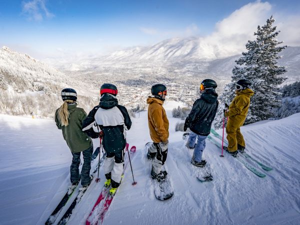 Five people with skis and snowboards stand on a snowy mountain slope, overlooking a scenic winter landscape.
