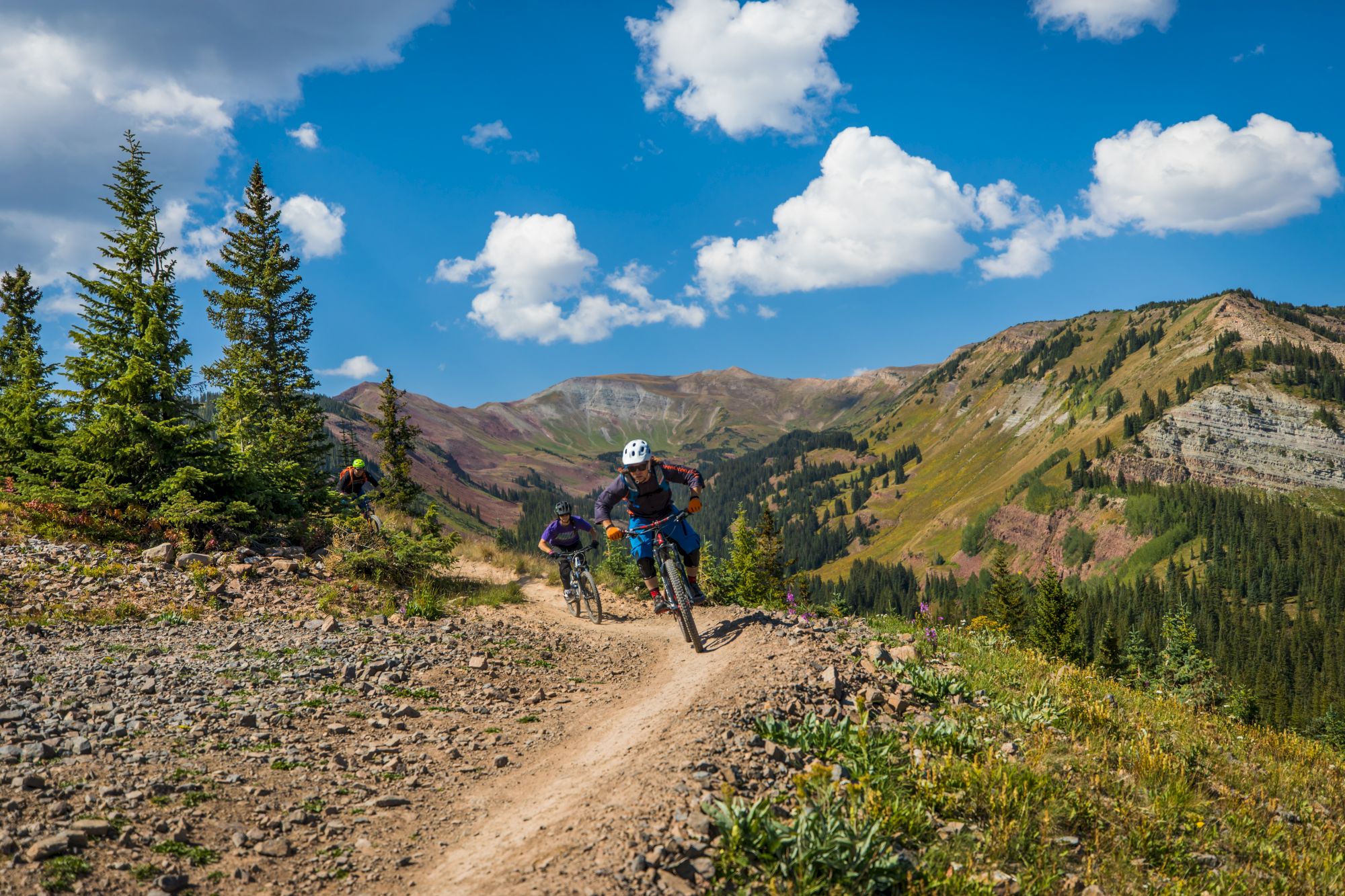 People are mountain biking on a rugged trail with scenic views of mountains, trees, and a blue sky with clouds in the background.